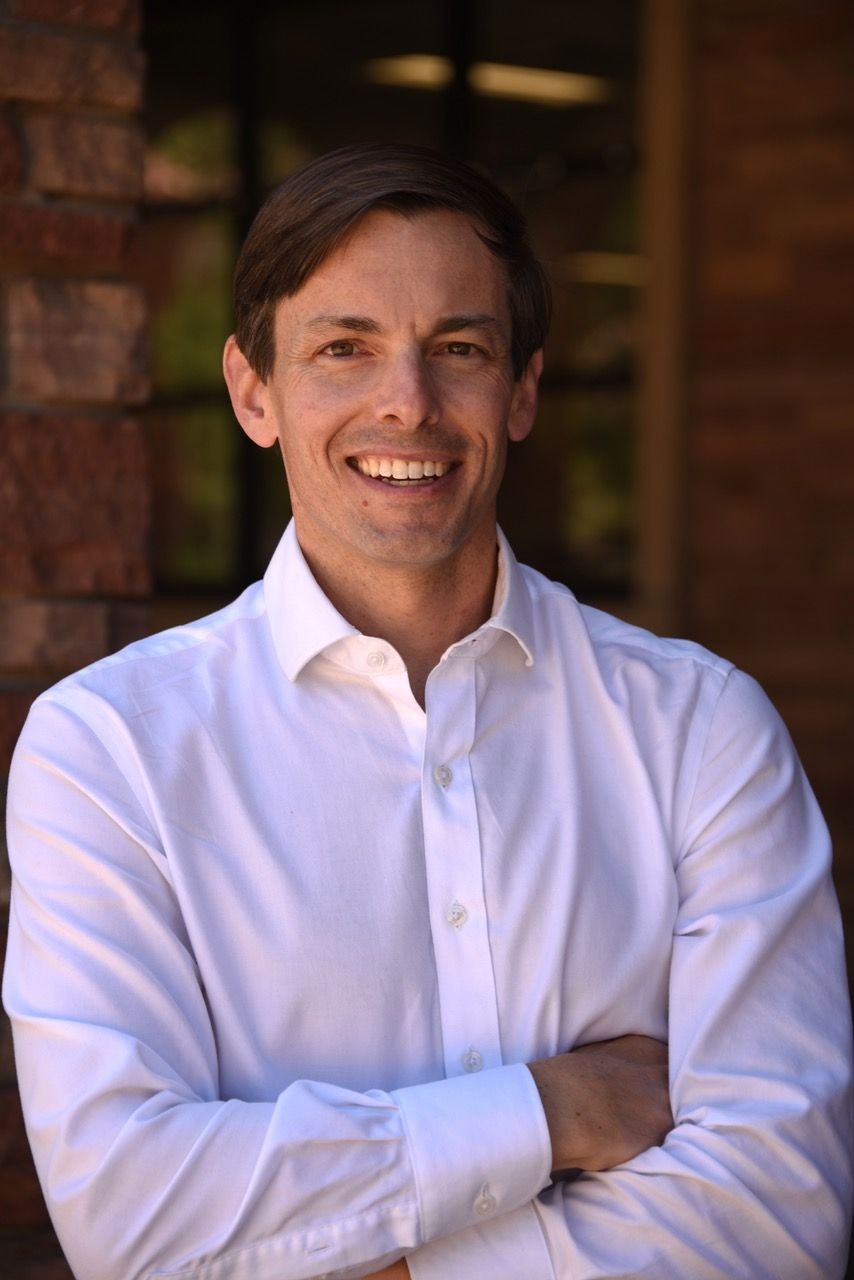 Person wearing a white dress shirt, standing in front of a brick wall with arms crossed