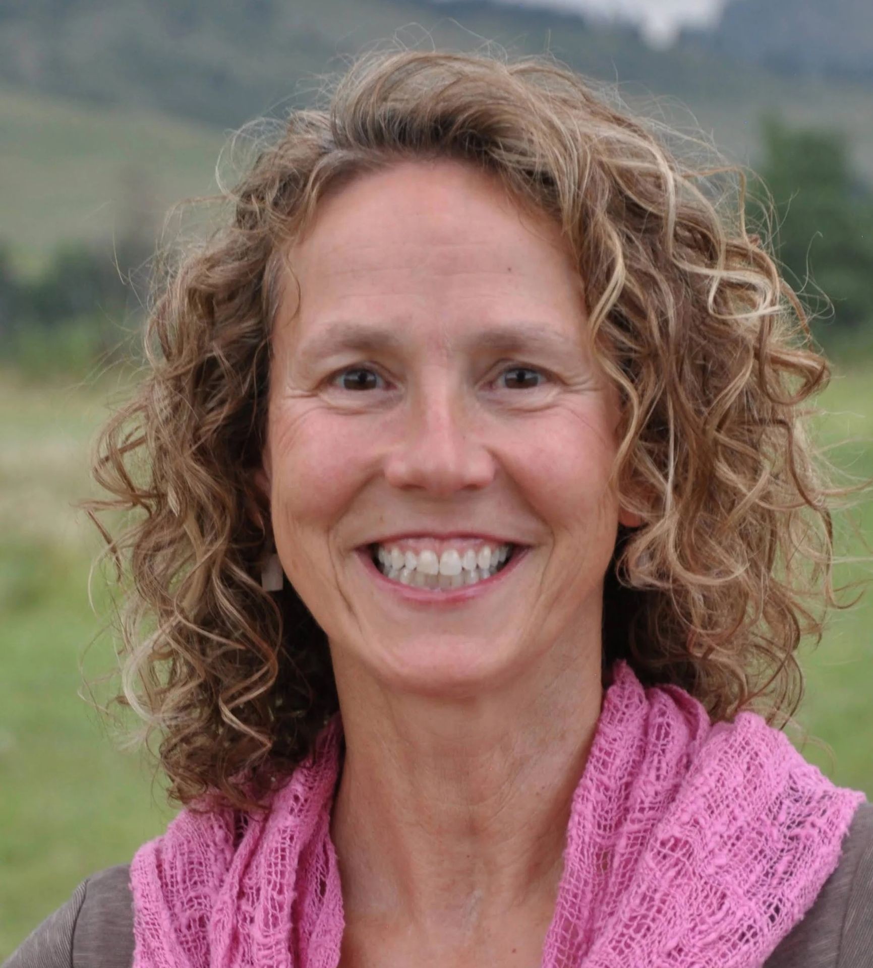 Smiling woman with curly hair wearing a pink scarf, standing outdoors.