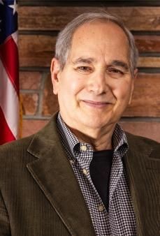 Smiling man in a brown jacket with an American flag in the background.