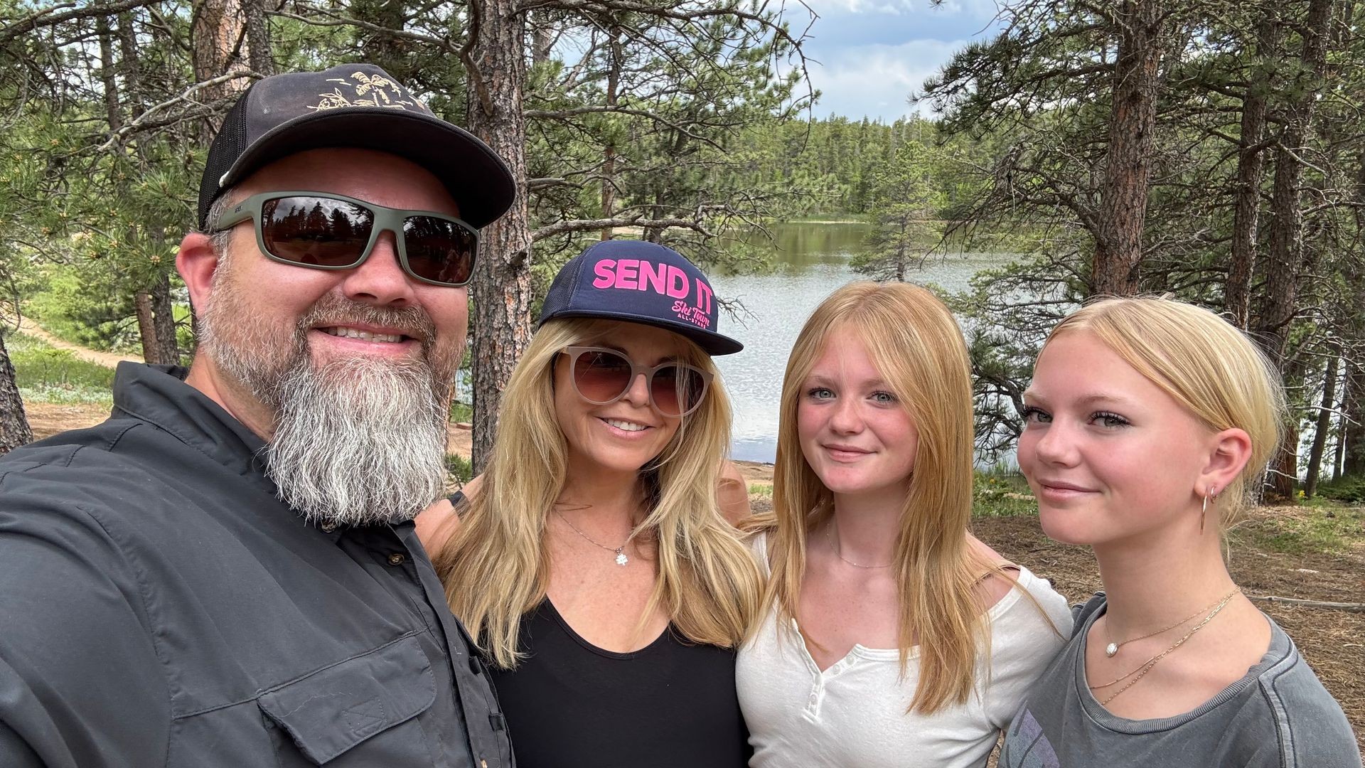 Group of four people standing outdoors surrounded by forest trees and a lake in the background.