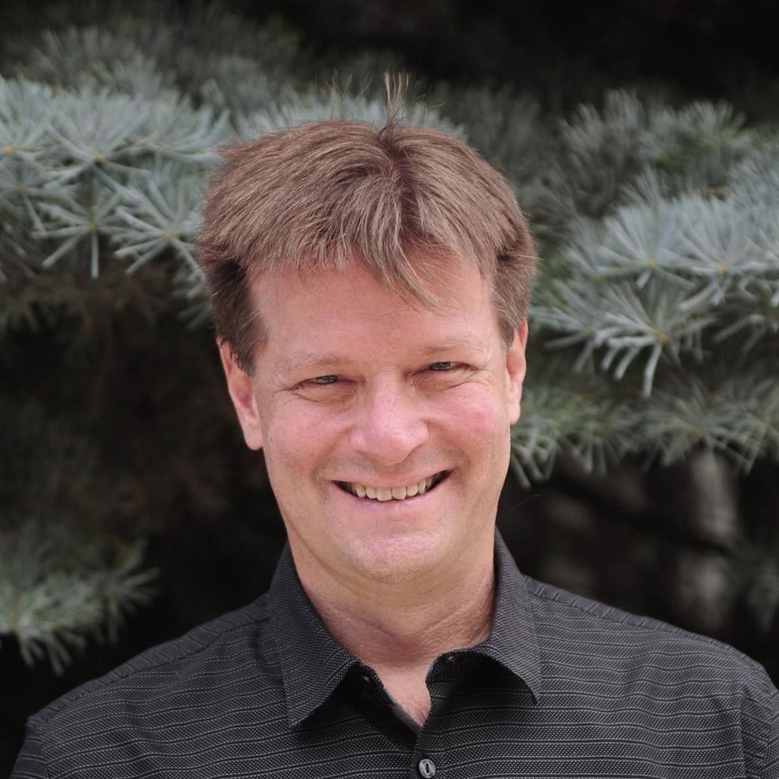 Man with brown hair wearing a dark grey shirt standing in front of a pine tree background.