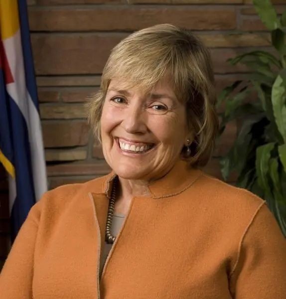 Smiling person wearing an orange jacket in front of a flag and plant.