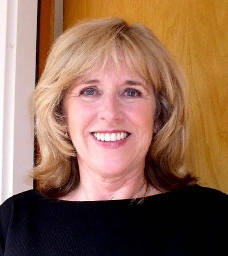 Smiling person with blonde hair wearing a black top, standing indoors against a wooden door.