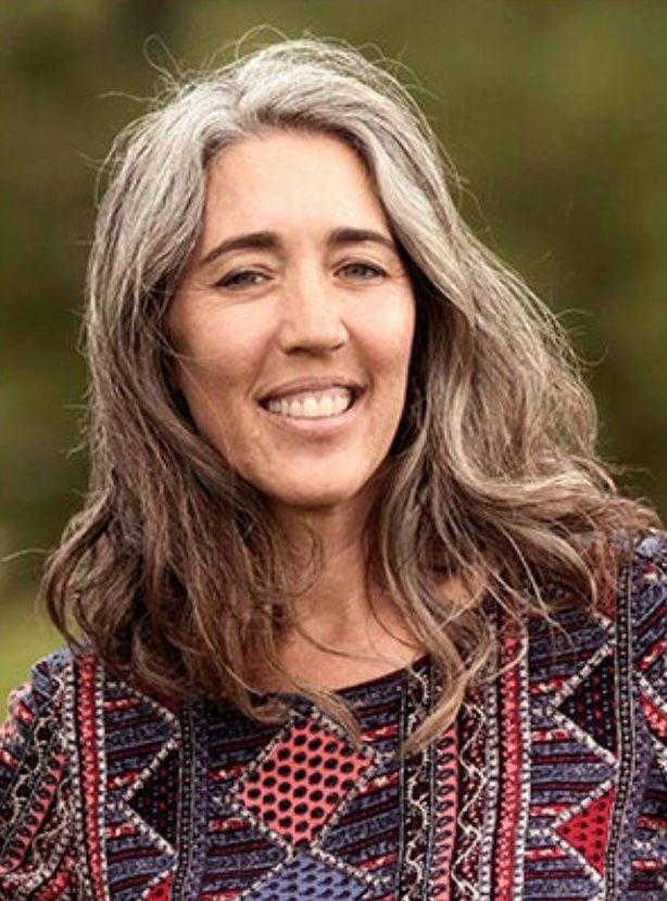 Smiling woman with long gray hair wearing a patterned top, outdoors.