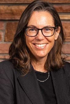Person with glasses and a black blazer smiling in front of a brick wall background.