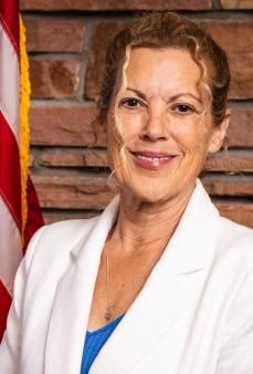 Smiling woman in white blazer poses in front of brick wall with American flag.