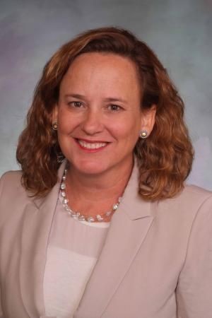 Woman with curly hair wearing a light-colored blazer and pearl necklace smiling against a neutral background.