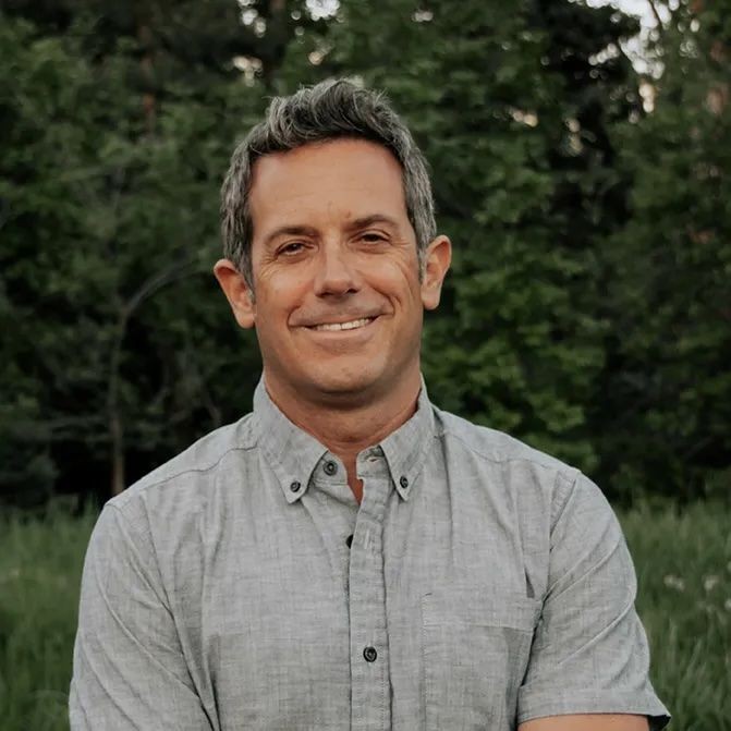 Man standing outdoors wearing a light grey button-up shirt, with a background of lush green trees.